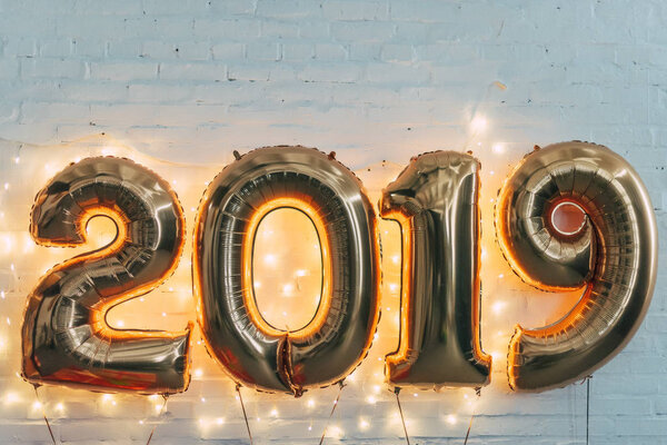 Золотые воздушные шары 2019 года со светлой гирляндой на белой стене к Новому году
