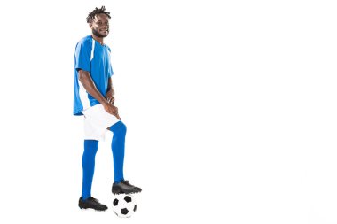 futbol topuyla duran ve üzerinde beyaz izole kamera gülümseyen atletik Afrikalı-Amerikalı sporcu görünümünü tam uzunlukta