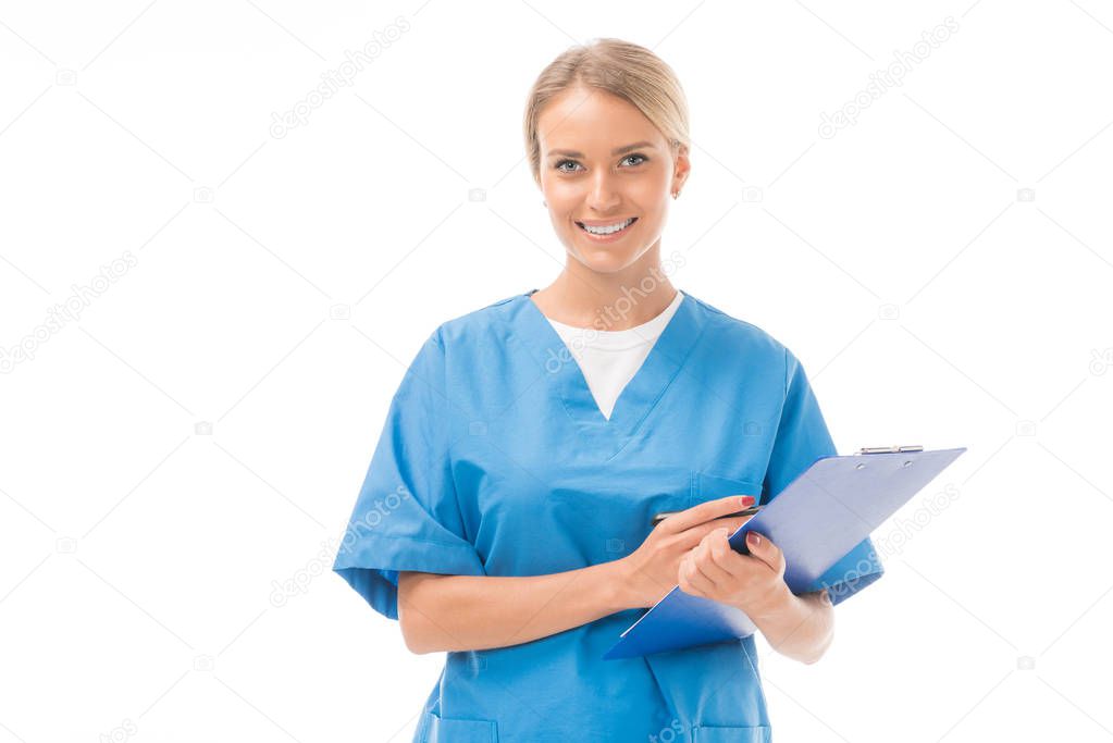 smiling young nurse writing in clipboard and looking at camera isolated on white