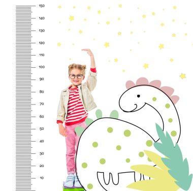 smiling child standing on pile of books to be higher isolated on white with imaginary dinosaur and growth measures clipart