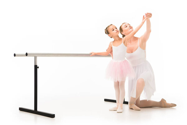 happy female teacher in tutu helping little ballerina exercising at ballet barre stand isolated on white background 