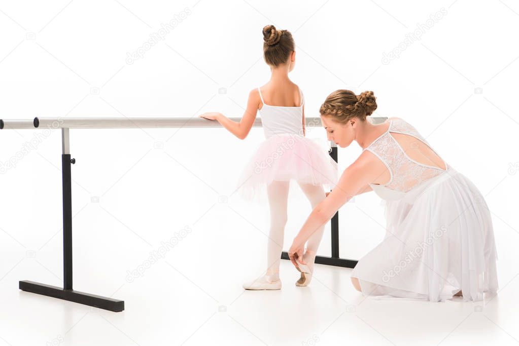 rear view of female teacher in tutu checking pointe shoes of little ballerina exercising at ballet barre stand isolated on white background 