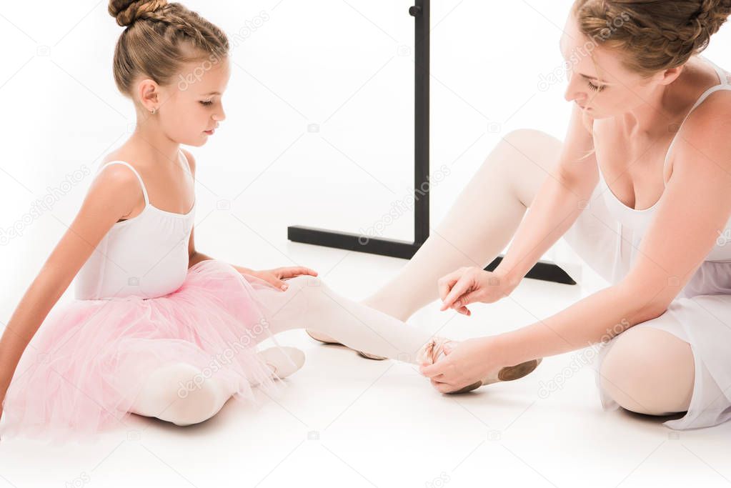 adult female trainer pointing at pointe shoes of little ballerina near ballet barre stand isolated on white background 