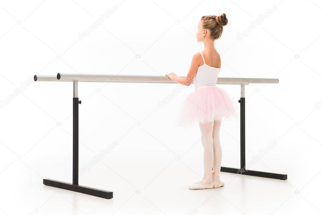 rear view of little ballerina in tutu and pointe shoes practicing at ballet barre stand isolated on white background 