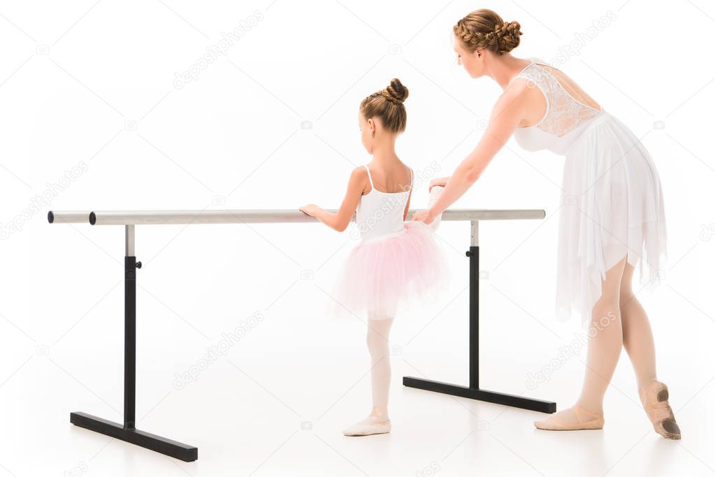 rear view of adult female teacher in tutu helping little ballerina exercising at ballet barre stand isolated on white background 