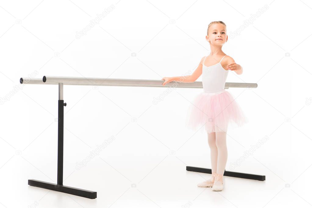 adorable little ballerina in tutu and pointe shoes practicing at ballet barre stand isolated on white background 