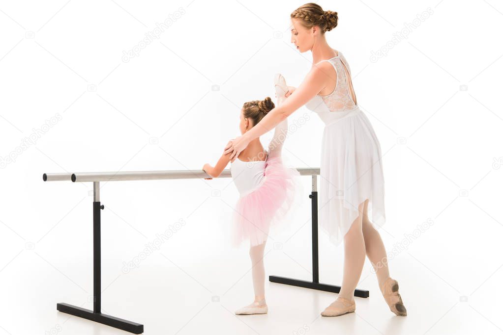side view of female teacher in tutu helping little ballerina practicing at ballet barre stand isolated on white background 