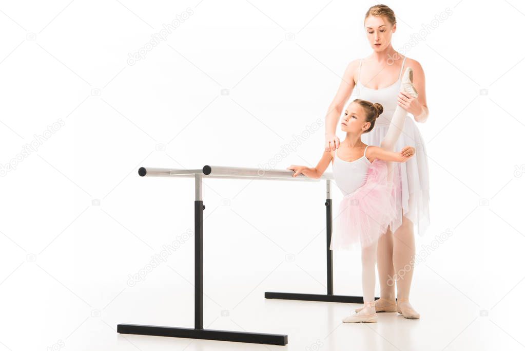focused female teacher in tutu helping little ballerina practicing at ballet barre stand isolated on white background 