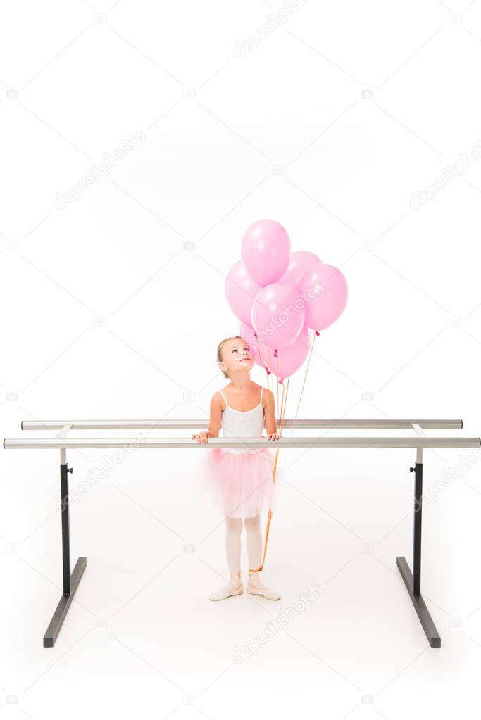 little ballerina in tutu standing at ballet barre stand and looking up at pink balloons isolated on white background 