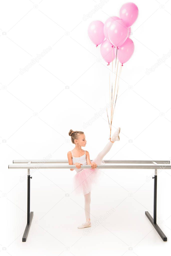 side view of little ballerina in tutu practicing with pink balloons wrapped over leg at ballet barre stand isolated on white background 