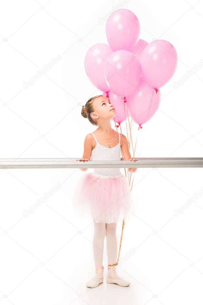 beautiful little ballerina in tutu standing at ballet barre stand and looking up at pink balloons isolated on white background 