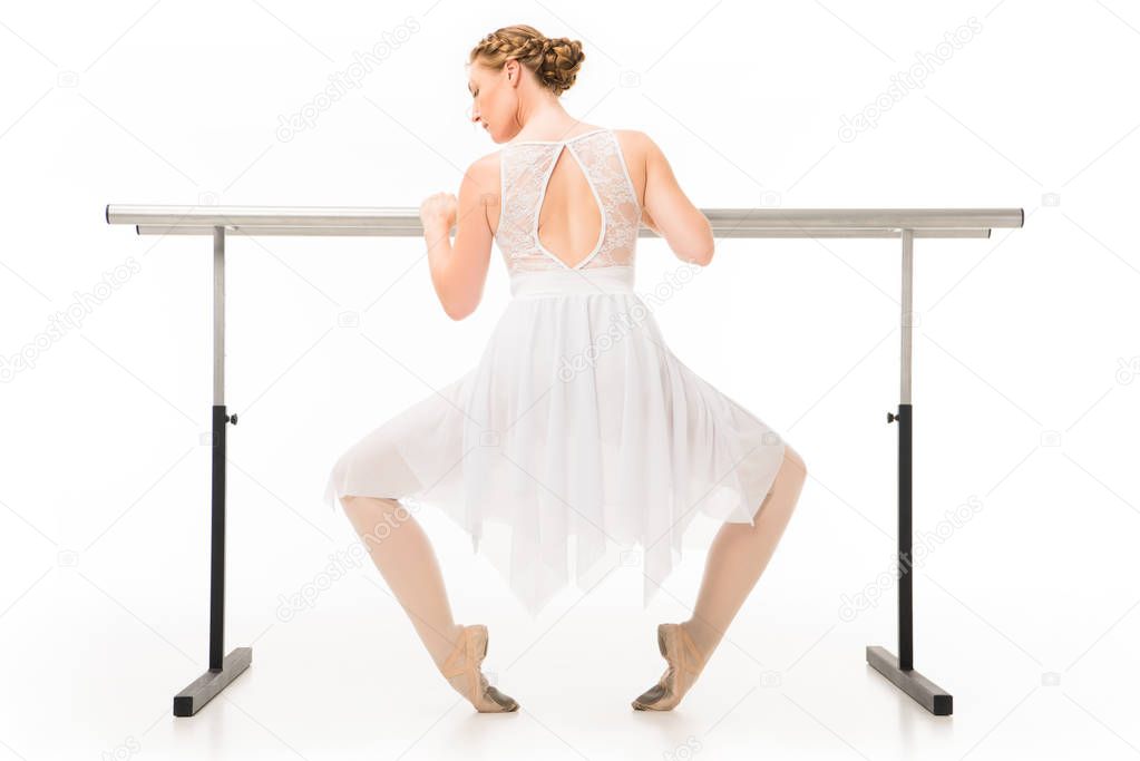 rear view of attractive ballerina in tutu exercising at ballet barre stand isolated on white background 