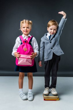adorable schoolboy standing on books to be higher near smiling schoolgirl with pink backpack clipart