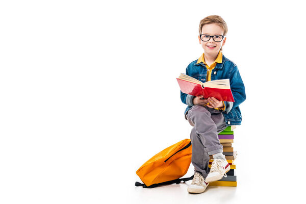 cheerful schoolboy reading book and sitting on pile of books with backpack, isolated on white 