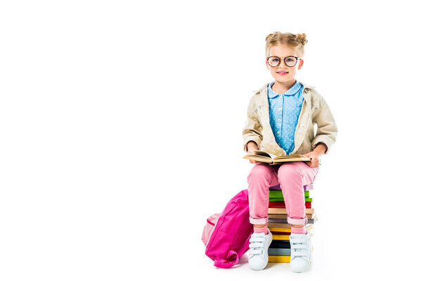 adorable pupil reading while sitting on pile of books with pink backpack isolated on white