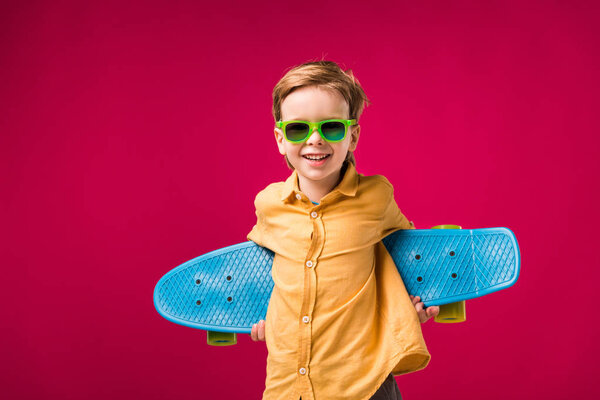 stylish cheerful boy in sunglasses posing with penny board isolated on red