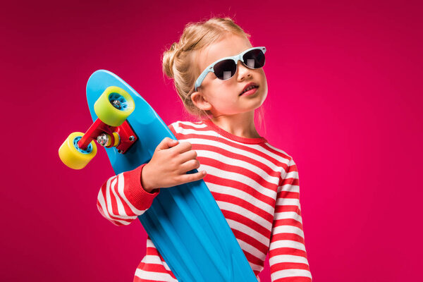 stylish kid in sunglasses posing with skateboard isolated on red