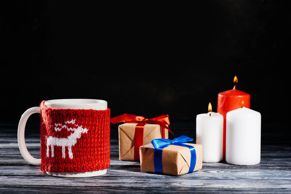 close-up view of cup, burning candles and christmas gifts on wooden table on black
