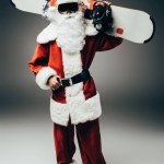 Babbo Natale in maschera da sci tenendo lo snowboard sopra la spalla su sfondo grigio