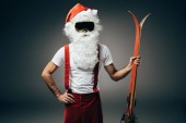 ernsthafter Weihnachtsmann in Skimaske, der mit der Hand auf der Taille steht und die Skier isoliert auf grauem Hintergrund hält 