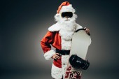 selbstbewusster Weihnachtsmann in Skimaske, der mit der Hand auf der Taille steht und das Snowboard isoliert auf grauem Hintergrund hält 