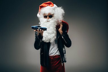 Noel Baba güneş gözlüğü ve deri ceket gri arka plan üzerinde izole oyuncak araba ve Noel çuval holding 