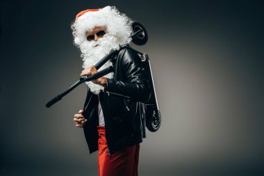 güneş gözlüğü ve deri ceket gri arka plan üzerinde izole omuz üzerinde tekme scooter tutan şık Noel Baba 