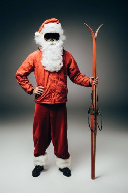 kar maskesi ve bel üzerinde el ile ayakta ve gri arka plan üzerinde kayaklar tutan rüzgarlık ciddi santa claus 
