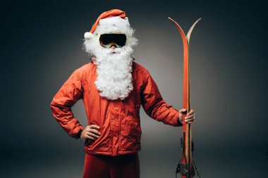 emin Noel Baba'ya kar maskesi ve bel üzerinde el ile ayakta ve gri arka plan üzerinde izole kayaklar tutan rüzgarlık 