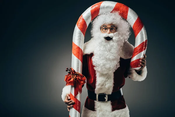 惊奇圣诞老人在服装站立与大条纹圣诞节棍子隔绝在灰色背景上 — 图库照片