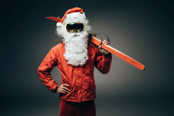 圣诞老人在滑雪面具和风衣举行滑雪板在肩膀和站立与手在腰部被隔绝的灰色背景 — 图库照片