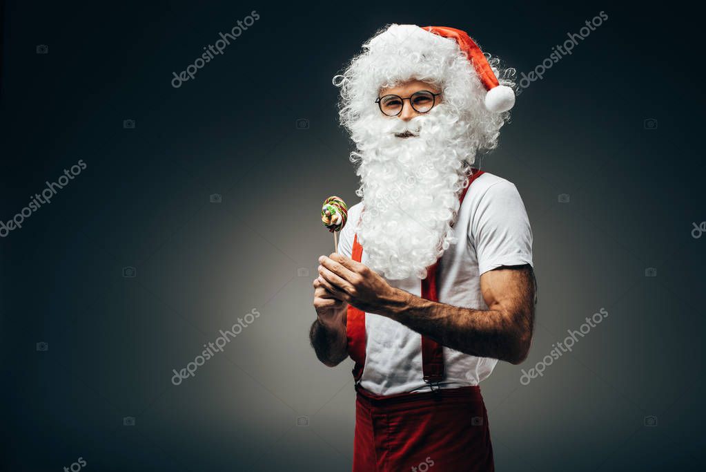 Happy santa claus in eyeglasses standing with colorful lollipop isolated on grey background