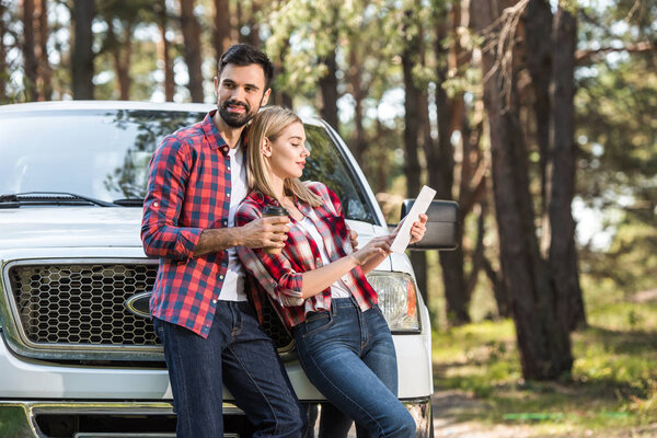handsome young man with coffee cup standing near girlfriend while she using digital tablet near pick up car outdoors 