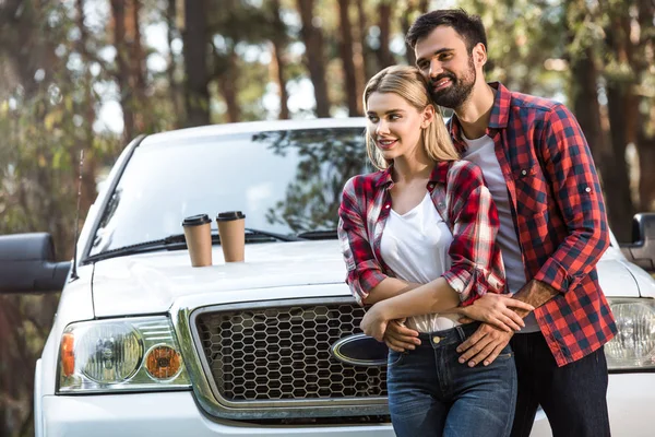 微笑的年轻人拥抱女朋友附近拾起汽车与咖啡杯子在帽子室外 — 免费的图库照片