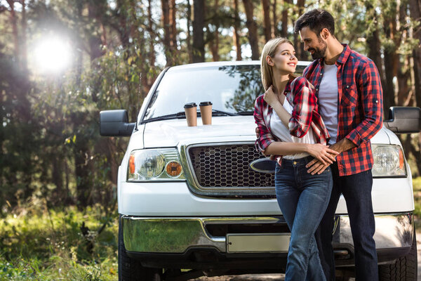 handsome man embracing girlfriend near pick up car with coffee cups on bonnet outdoors 
