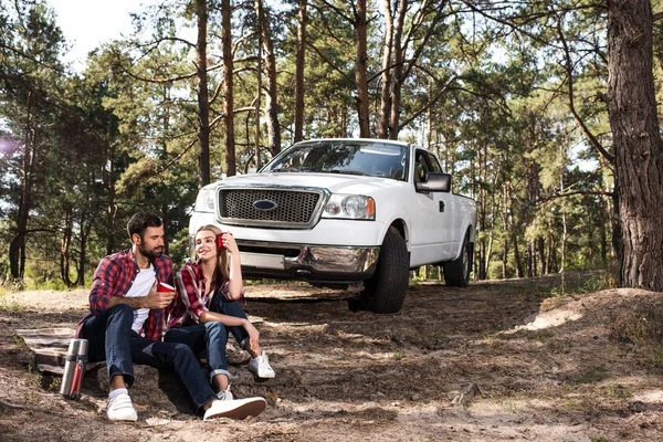 そばにはコーヒー カップと地面に座って若いカップルが森で車をピックアップします — ストック写真