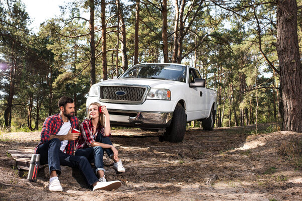 young couple sitting on ground with coffee cups near pick up car in forest