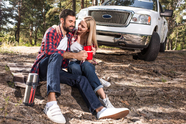 cheerful couple sitting on ground and clinking by coffee cups near pick up car in forest
