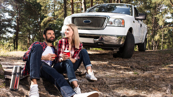 smiling young couple sitting on ground with coffee cups near pick up car in forest 