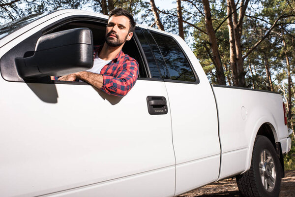 serious young handsome man sitting in pick up car and looking at wing mirror in forest 