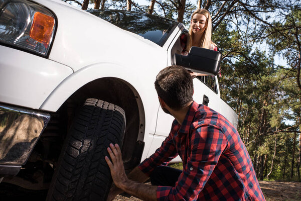 rear view of man fixing wheel while his girlfriend sitting inside of pick up car outdoors 