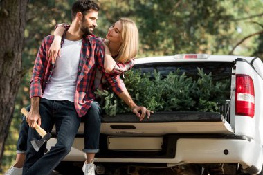 handsome young man holding axe and standing near girlfriend sitting in car trunk with christmas tree in forest clipart