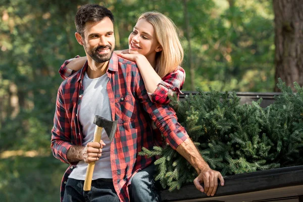 陽気な若い男が斧を押しながら森でクリスマス ツリーと車のトランクに座ってのガール フレンドの近くに立って — ストック写真