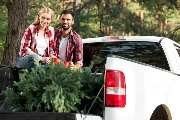 微笑的夫妇与杯子坐在汽车树干与圣诞树户外 — 图库照片