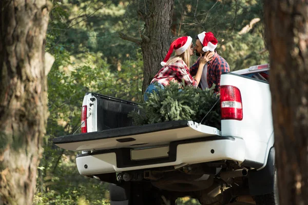 两口子戴着圣诞礼帽在汽车行李箱里接吻 森林里挂着圣诞树的低视角照片 — 图库照片