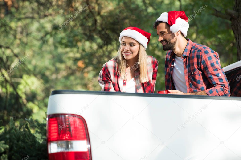 smiling young couple in santa hats with cups sitting in car trunk in forest 