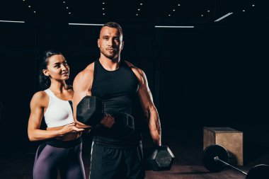 Barbell ile egzersiz ve kamera arkasında koyu spor salonunda duran kadın süre bakarak atletik vücut geliştirmeci