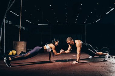 athletic sportsman and sportswoman doing push ups together and holding hands in dark gym clipart