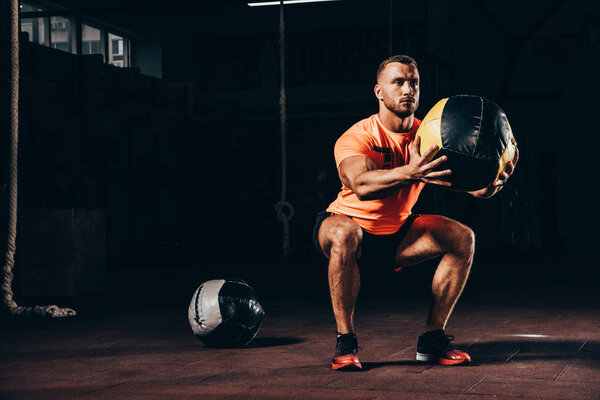 handsome athletic sportsman performing squats with medicine ball in dark gym