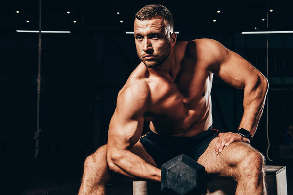 handsome athletic bodybuilder lifting dumbbell while sitting on cube in dark gym